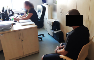 Policjantka siedzi przy biurku, przy komputerze, a obok niej zatrzymany mężczyzna. Na dłoniach ma założone kajdanki