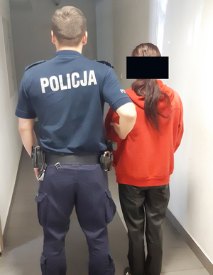 Policjant trzyma za ramię kobietę. Oboje stoją na korytarzu.