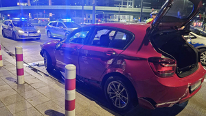 Czerwony samochód zaparkowany na ulicy z widocznymi wgnieceniami po uderzeniu, a obok stoją dwa radiowozy policyjne.