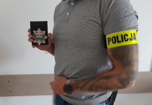 Mężczyzna  w dłoni, trzyma odznakę policyjna, a na przedramieniu ma założoną opaskę z napisem Policja.