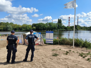 Dwóch policjantów dzielnicowych stoją na plaży w pobliżu kąpieliska wodnego.