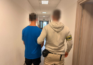 Policjant z osobą zatrzymaną stoją tyłem. Zatrzymany na rękach ma założone kajdanki